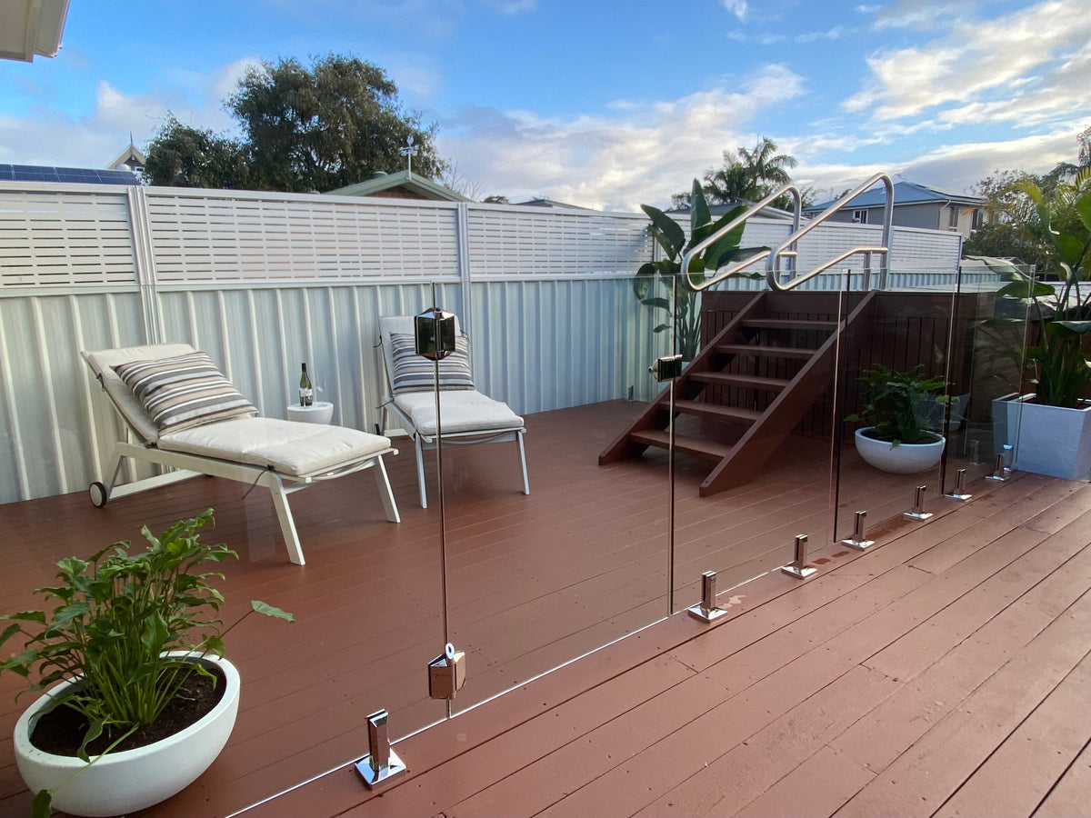 A modern glass railing system with stainless steel posts and handrails installed on a reddish-brown wooden deck, shown in an outdoor backyard setting with lounge chairs, a staircase, and plants. A White colorbond fence and a white slat Screenline fence extension.
